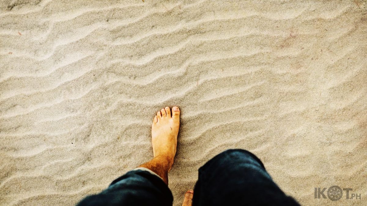 A man in folded blue denim jeans walking through the sand.